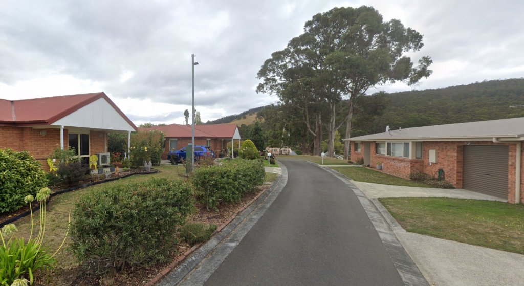 A narrow, paved street curves between single-story brick houses with well-kept lawns and gardens, surrounded by green hills and tall trees under a cloudy sky.