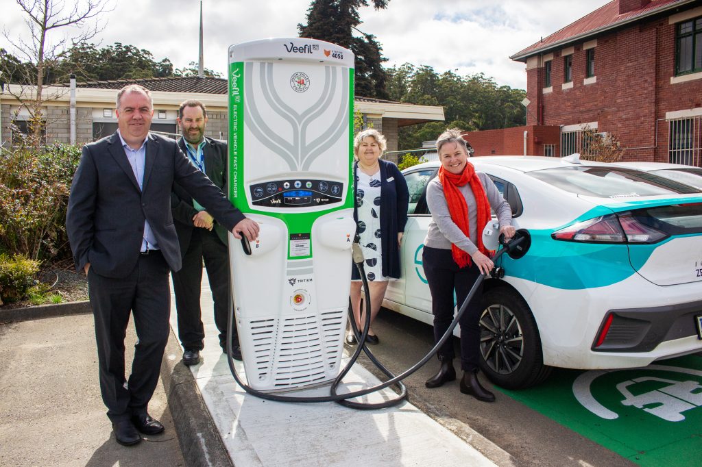 Four people stand around an electric vehicle charging station. One person is plugging a white electric car into the charger. The station is located in a parking area with buildings and trees in the background.