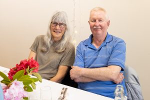 An elderly couple is sitting at a table against a plain wall. The woman has long gray hair and glasses, wearing a gray T-shirt. The man has short gray hair, a mustache, and is wearing a blue striped polo. Bright flowers are on the table in front of them.