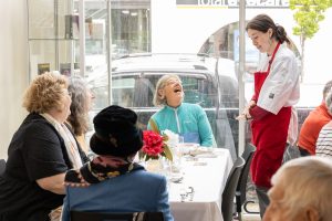 A waitress in a red apron talks to a group of four smiling elderly women seated at a table in a cafe. One woman is laughing heartily. The table has a white tablecloth and a vase with red flowers.
