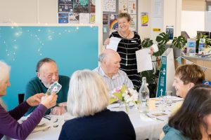 A group of elderly people sitting around a table, smiling and looking at cards. A woman stands smiling, holding papers. The setting has a turquoise backdrop and is decorated with flyers and string lights. There are flowers and water bottles on the table.