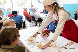 A waitress in a red apron and white chefs hat serves a dish to a seated patron at a busy restaurant. Other diners are visible in the background, sitting at tables adorned with white tablecloths and pink flowers.
