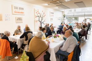 A group of people seated around a long table in a bright room, enjoying a meal together. Walls display artworks and banners. Natural light streams in through large windows, creating a warm and inviting atmosphere.