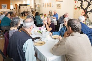 A group of older adults seated around tables covered with white tablecloths, engaged in conversation and laughter. The room is decorated with art on the walls and flower arrangements on the tables.