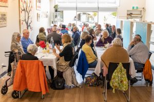 A group of people of various ages are sitting at tables in a bright, open room with large windows. They are engaged in conversation and dining, with decorations and artwork on the walls.