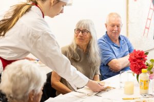 A young person in a chefs uniform serves food to three older adults seated at a table. One woman with glasses smiles up at the server, while a man in a blue shirt sits next to her. The table is decorated with red flowers and candles.