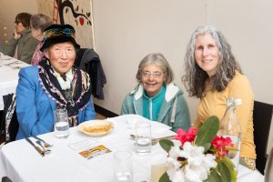 Three women sitting at a dining table with white cloth, smiling at the camera. There are plates and glasses on the table, along with a vase of vibrant flowers. A decorative artwork is partially visible on the wall behind them.