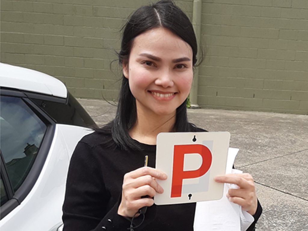 A person smiling and holding a sign with a red P on it, standing in front of a white car. The person is wearing a black top and has long, dark hair. A green wall is in the background.