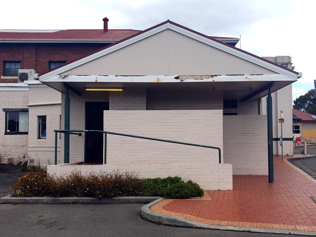 A small building with a sloped roof, light brick walls, and a red brick pathway. It has a green handrail along a ramp. The roofs edge shows some wear. An air conditioning unit is visible on the side. The sky is overcast.