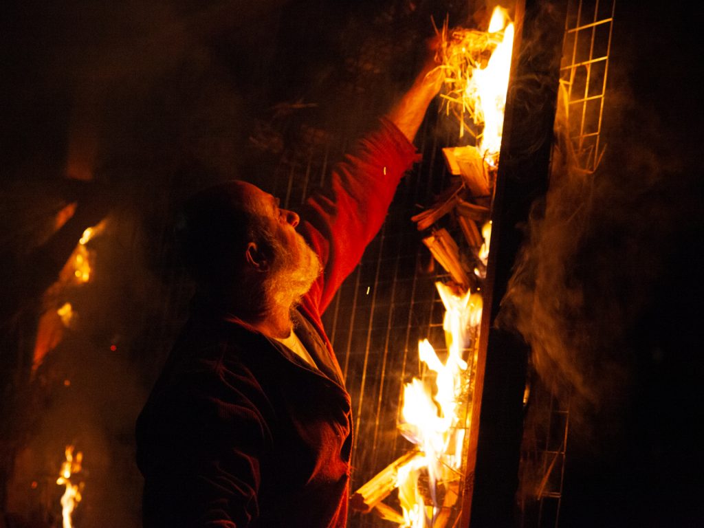 A person in a red jacket places a bundle of kindling onto a burning fence at night. The flames illuminate the scene with a warm glow, and smoke rises around the person, creating a dramatic and intense atmosphere.