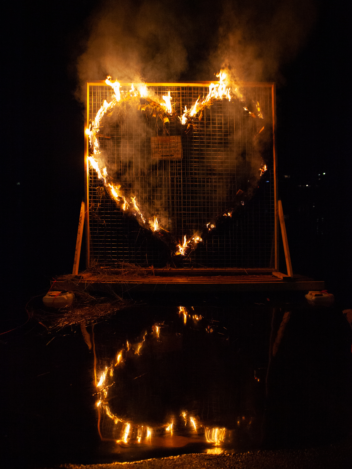 A large heart-shaped structure is on fire, surrounded by a metal grid. The fire illuminates the night, casting a glowing reflection on the water below. Smoke rises into the dark sky, adding a dramatic effect.
