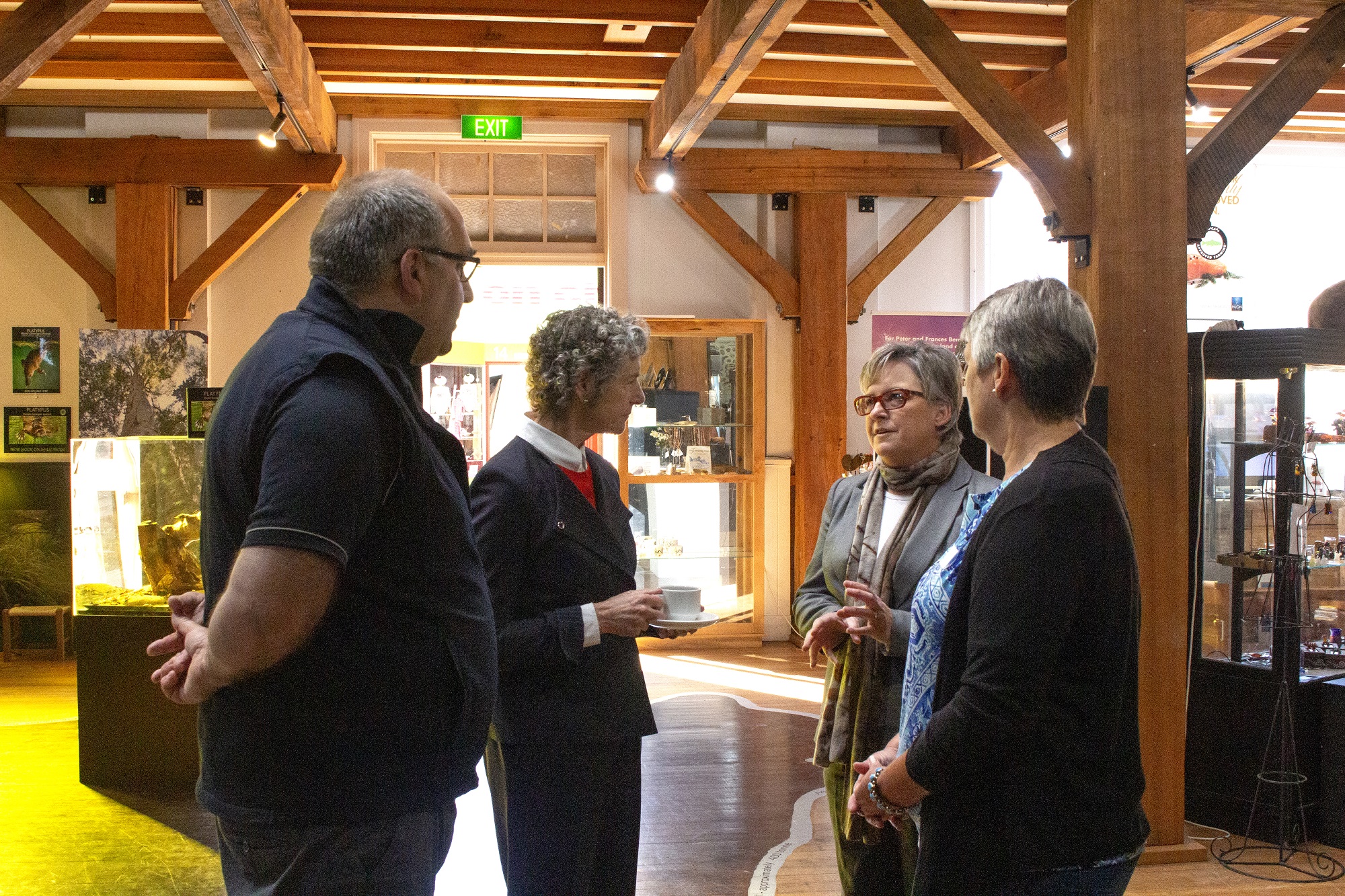 Four people stand in a warmly lit room with wooden beams, engaged in conversation. Various exhibits and framed pictures are displayed in the background.