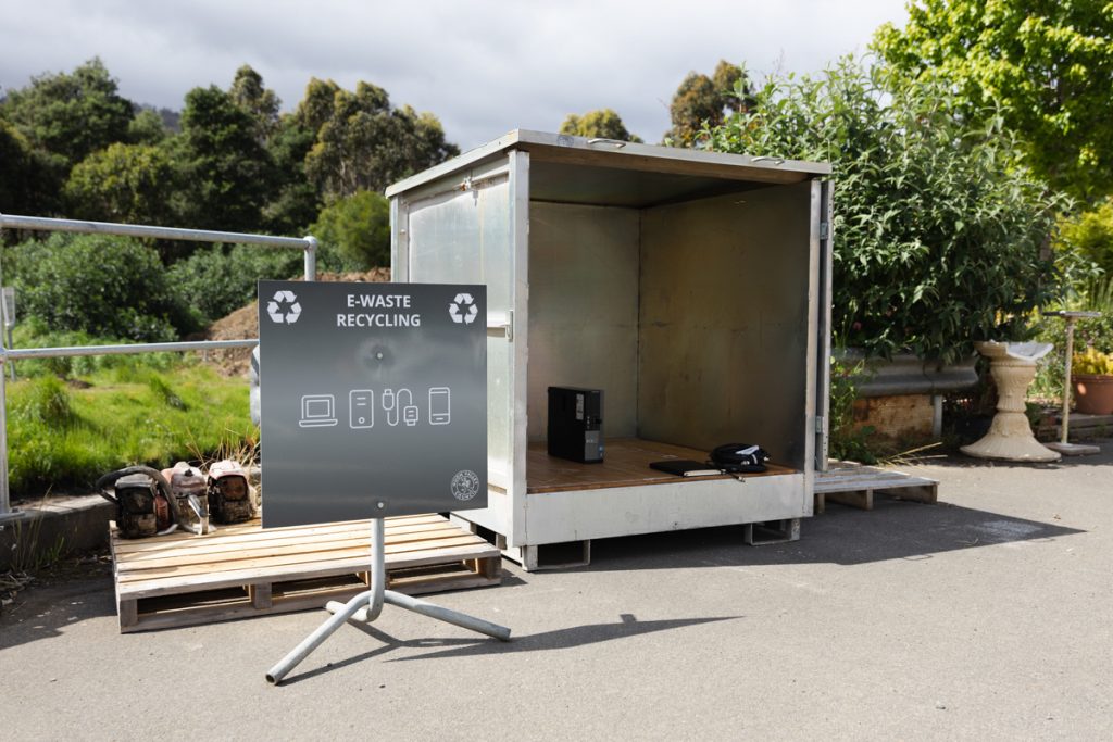 An outdoor e-waste recycling station with a metal container on wooden pallets. A sign nearby displays icons for electronic items like phones and computers. Trees and a cloudy sky are in the background.