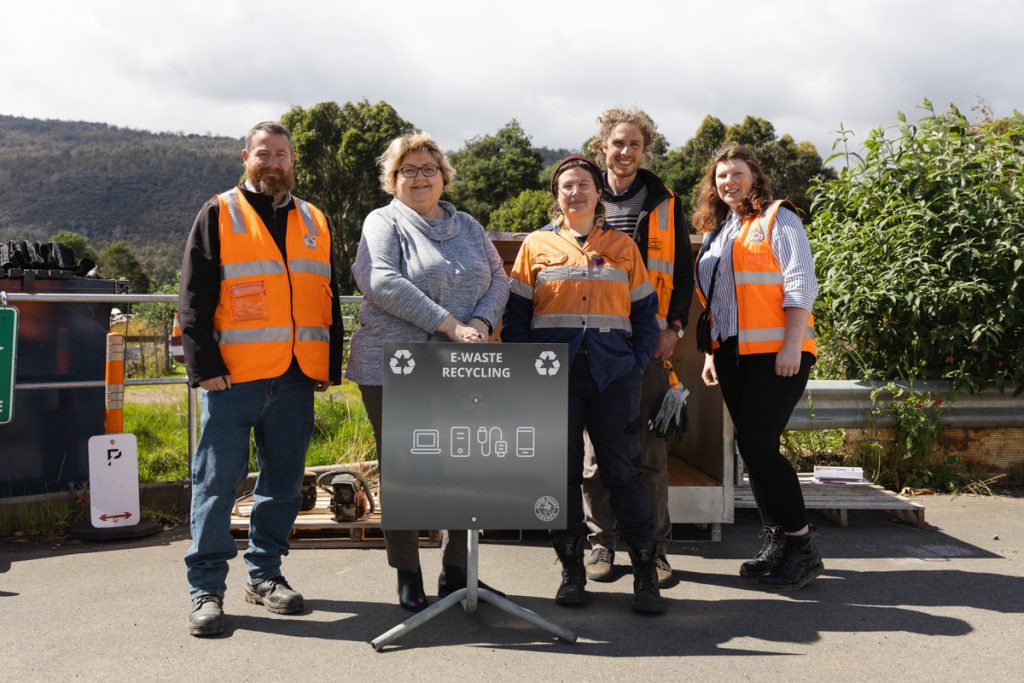 Five people in reflective vests stand outdoors around a sign that reads E-Waste Recycling. They are smiling at the camera, with mountains and trees visible in the background. Various electronic items are scattered nearby.