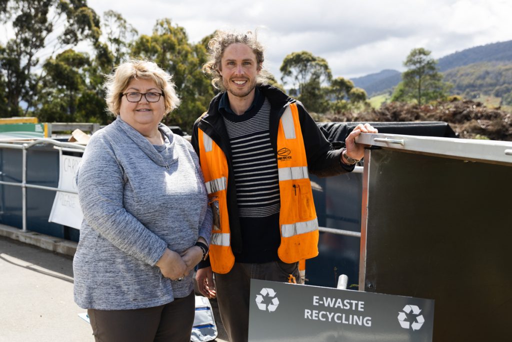 Two people stand outdoors near e-waste recycling bins. The woman on the left is wearing glasses and a gray sweater. The man on the right is wearing an orange safety vest over a striped shirt. Trees and hills are visible in the background.