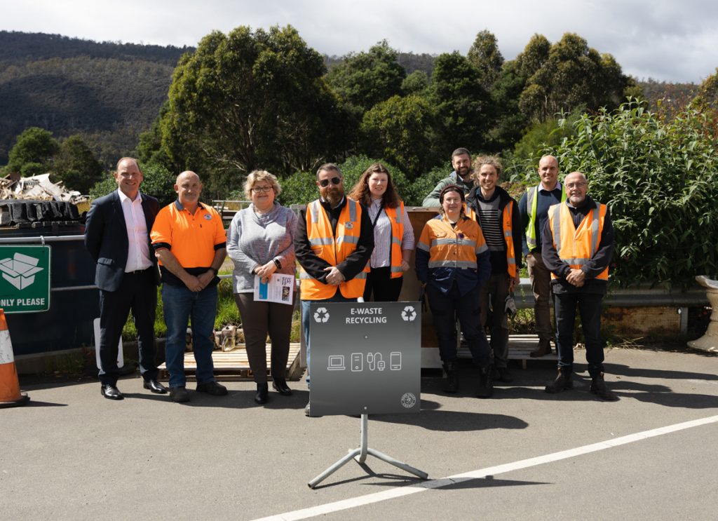 A group of people, some in orange safety vests, stand outdoors near recycling bins and a sign labeled E-waste Recycling. Trees and a hill are visible in the background. They appear to be posing for a photo.
