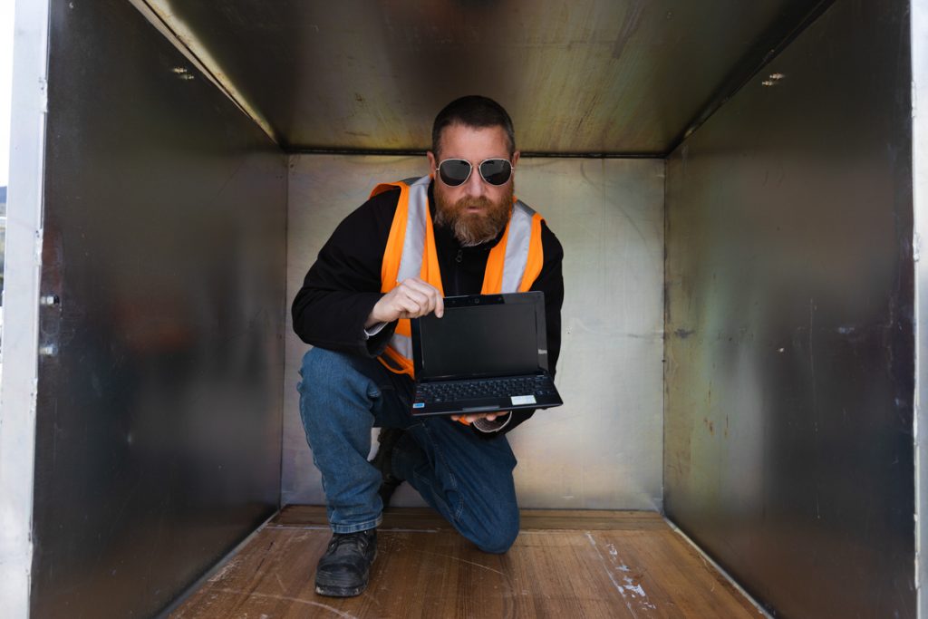 A bearded man wearing sunglasses and an orange safety vest crouches inside a large metal container. He is holding an open laptop, displaying the screen towards the viewer.