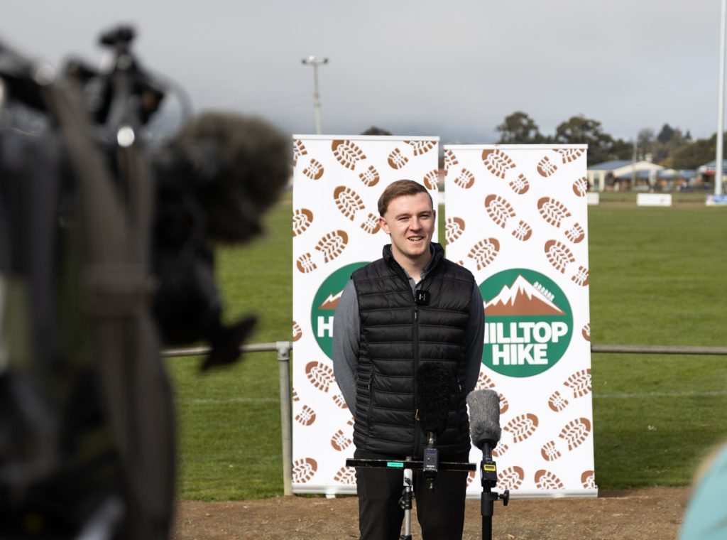 Young man in a black jacket stands in front of microphones, giving an outdoor speech. Behind him are banners with a logo that reads Hilltop Hike, featuring mountain and footprint graphics. A blurred camera is in the foreground.