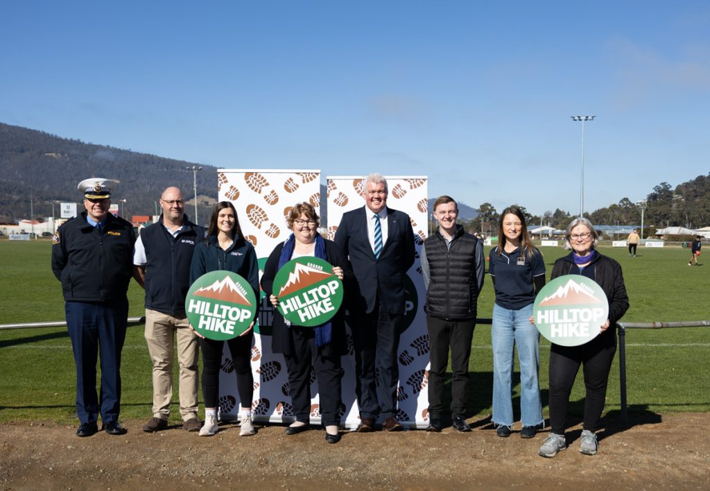 A group of people, including uniformed personnel and individuals holding Hilltop Hike signs, stand together on a sports field. A banner with logos is behind them, and distant mountains are visible under a clear blue sky.