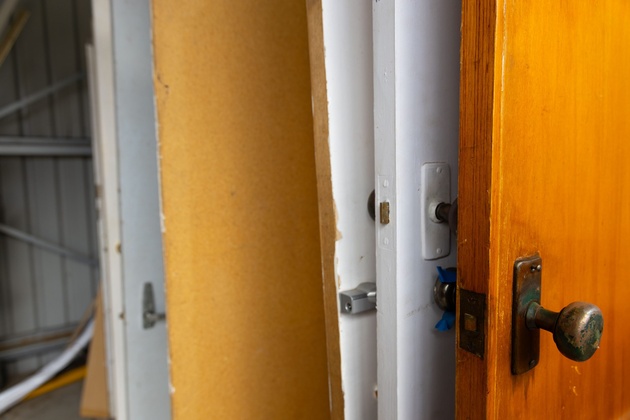 A row of standing doors in a shed. The closest is a wooden door with a dark brass doorknob. Behind it are several other doors, in varying colors and conditions, leaning against a wall. The sheds metal structure is visible in the background.