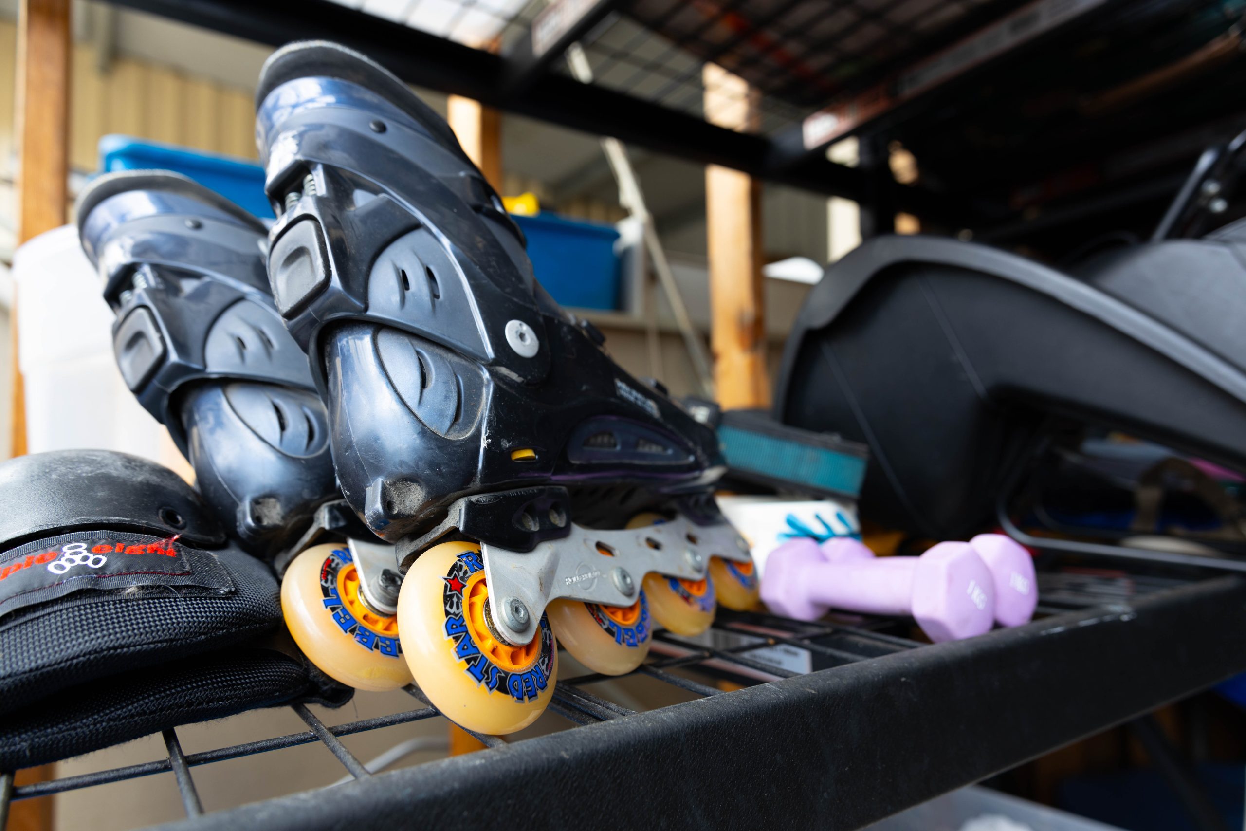 Close-up of rollerblades with orange wheels on a metal shelf. Nearby, there are purple dumbbells and protective gear, suggesting a focus on fitness and sports equipment.