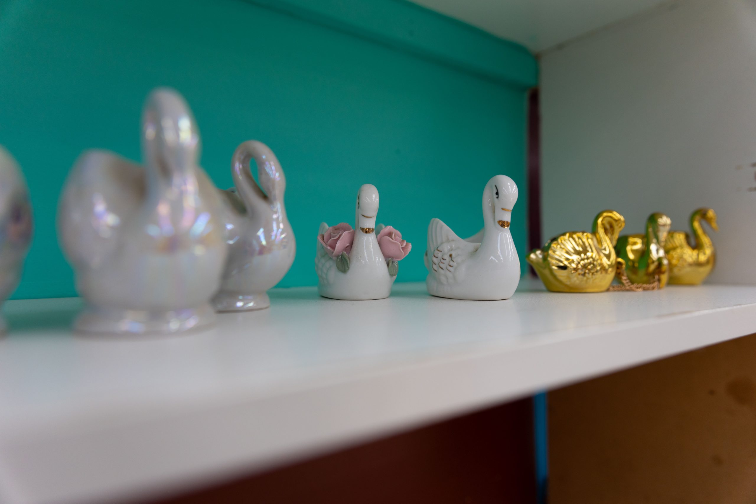 A shelf displaying swan figurines, including iridescent, white with pink flowers, and gold ones, set against a teal background.