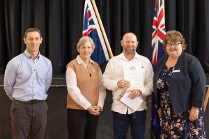 Four people are standing together in front of two Australian flags. One person is holding a certificate. They are all dressed in semi-formal attire and smiling. There is a dark curtain in the background.