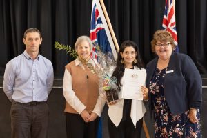 Four people stand together indoors in front of Australian flags. A woman holds a certificate and wrapped gift. They are dressed in formal attire and appear to be at a ceremony.