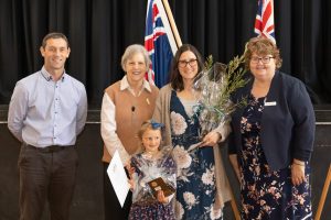 A group of five people, including two children, stand together smiling. One holds a certificate, and another holds a small plant. Two flags are in the background. The setting appears to be an indoor event or ceremony.