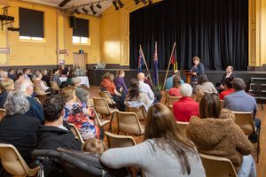A woman speaks at a podium in a community hall. She faces an audience sitting in rows of chairs. Behind her, there are three flags and a black stage curtain. The audience includes adults, children, and a person with a stroller.