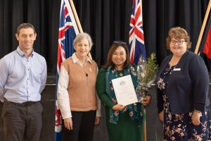 Four people standing and smiling in front of flags. One woman holds a certificate and a potted plant. They are in a room with a black backdrop.