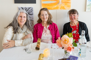 Three women are seated at a decorated table with flowers, a candle, and a bottle of water. They are smiling, wearing casual attire, and theres artwork on the wall behind them.