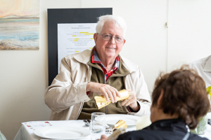 An older man with white hair and glasses, wearing a beige jacket over a red and green shirt, smiles while seated at a dining table. He is opening a yellow gift. Another person is partially visible in the foreground.
