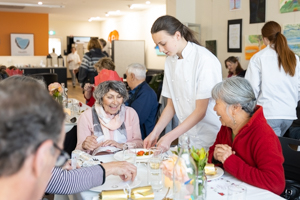 A young person in a white shirt serves food to two older adults sitting at a table in a restaurant. The diners are smiling and conversing, surrounded by other patrons and colorful decorations.