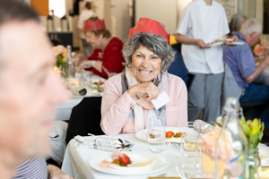 An elderly woman with gray hair, wearing a red paper crown, smiles while sitting at a table with a white tablecloth and a plate of food. Other people are dining in the background.