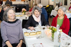 Three women are sitting at a table, smiling and laughing together at a social event. The table is set with plates, glasses, and floral centerpieces. Other attendees are visible in the background.