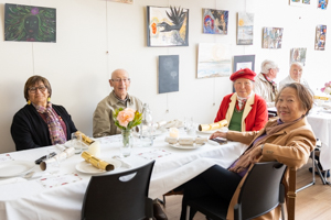 Four elderly individuals are seated around a table set with plates and napkins in a room decorated with paintings. One person wears a red hat. They appear to be in a casual dining setting, possibly an art gallery or a community space.