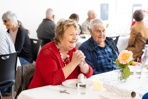 A smiling elderly woman in a red top and an elderly man in a plaid shirt sit at a table with plates and a rose centerpiece. Other people are seated at tables in the background, enjoying a meal together in a bright room.