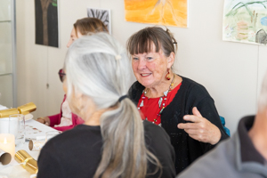 A group of people sitting around a table engaged in conversation. A woman with a red shirt and earrings is smiling and talking to another person with gray hair tied back. Artwork is visible on the wall in the background.