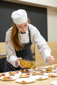 A chef wearing a white uniform and striped apron is plating dishes. She is focused and holding a bowl with a yellow food mixture, spooning portions onto several white plates arranged on a table.
