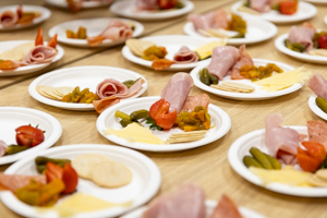 Plates with assorted appetizers, including rolled ham, cherry tomatoes, pickles, cheese slices, crackers, and diced vegetables, are arranged on a wooden table.