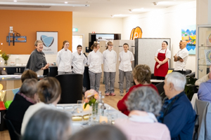 A group of chefs in white coats and gray pants stand in a line at the front of a room. A person in red apron stands with them. Seated audience members, some with gray hair, are listening. The room has a warm, informal setting.