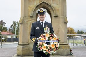 A uniformed naval officer stands in front of a monument holding a wreath adorned with flowers and ribbons that read Lest we forget. The background features autumn trees and cloudy skies.