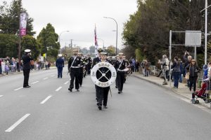 A marching band parades down a street, led by a drum major holding a bass drum with an emblem. The street is lined with spectators. Band members wear uniforms with caps, and one carries an American flag. Trees and a cloudy sky are in the background.