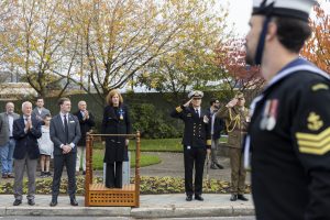 A woman stands on a platform saluting, surrounded by military personnel and civilians clapping. A sailor in the foreground is out of focus. Autumn trees and a flagpole are in the background.