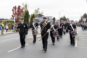 A marching band in formal uniforms parades down a street, led by a conductor with a staff. Several drummers and other musicians follow. Onlookers watch from the sidewalks, and autumn trees are visible in the background.