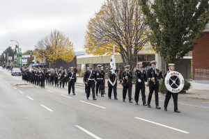 A marching band in uniform parades down a street lined with autumn trees. They hold instruments and a banner. Spectators watch from the sidewalk. Buildings and a parked car are visible in the background.