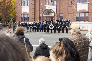 A group of uniformed individuals stands in front of a brick municipal building. People in the foreground are watching the group, and a tree with autumn foliage is visible to the left.