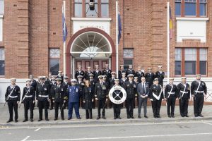 A group of people in uniform, including naval and police attire, stand in front of a brick municipal building. They pose with a drum in the center, some wearing medals and formal hats. Rows of flags are displayed on the building.