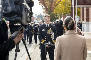 A naval officer in uniform is being interviewed on a street lined with people and trees with autumn foliage. A camera crew is filming the interaction. The officer stands by a microphone, facing a woman in a beige coat.